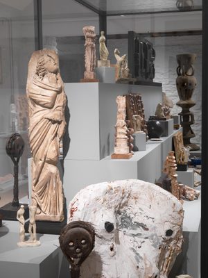 A large display case with a stepped plinth inside, holding numerous small sculptures by Henry Moore made in bronze, plaster and wood. They display a range of styles and techniques, from figurative sculptures to heads and more abstract pieces, and are accompanied by other sentimental objects.
