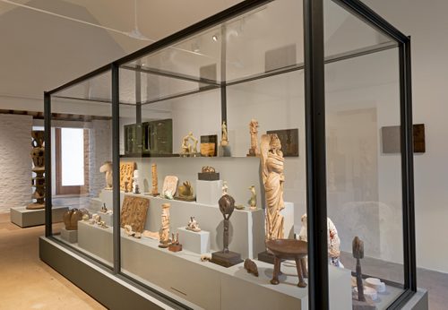 A large display case with a stepped plinth inside, holding numerous small sculptures by Henry Moore made in bronze, plaster and wood. They display a range of styles and techniques, from figurative sculptures to heads and more abstract pieces, and are accompanied by other sentimental objects.