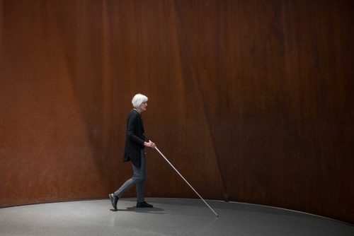 A woman with a cane walking past a wall made of rusted metal.