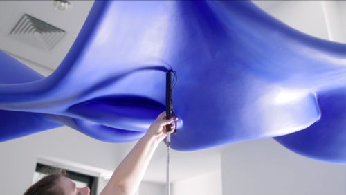 A person holding a cane reaches up to touch a large, blue, cloud-like fibreglass sculpture suspended from the ceiling.