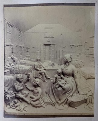 Black and white photo of a relief sculpture, possibly depicting a children's hospital. Beds with children in line the walls, with women tending to some of them. A man with a beard sits at a table in the centre. In the foreground, a woman holds one baby on her knee, while her outstretched hand touches another in a basket in the floor. Two children, one with its arm in a sling, play on the floor beside her.