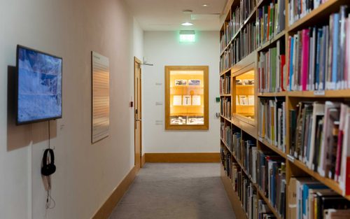 Corridor in a library, while shelves of books to the right, a screen on the left, and a display cabinet at the end.