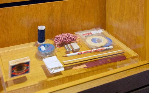 A display cabinet with stationary: a pencil, ruler, sharpener, eraser, masking tape, string, matches, and thread.