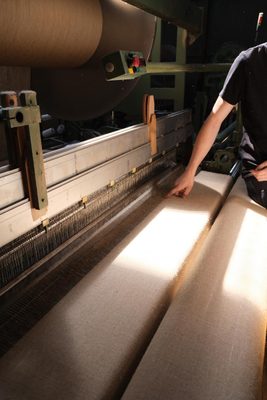 A mechanical loom weaving fibres into linen fabric. A man is checking the fibres with a finger as they pass through the loom.