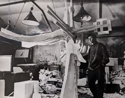 Black and white photo of a man in an art studio, looking at a large, cross shaped sculpture made from polystyrene.