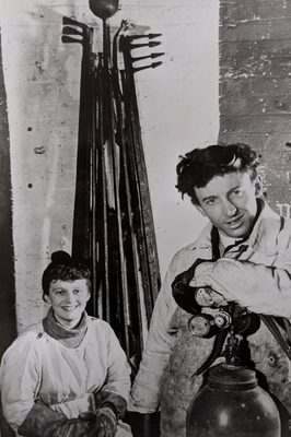 Photo of a man and a boy wearing heat resistant clothing, with a tall sculpture made of metal behind them.