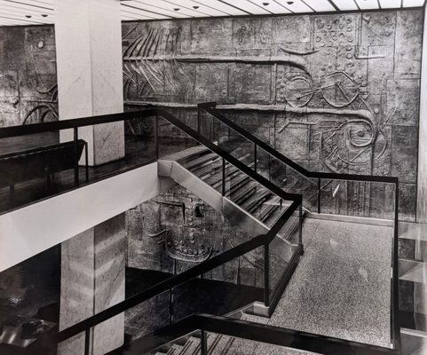 Black and white photo of a set of stairs, behind which is a large, abstract sculptural mural covering the entire wall.