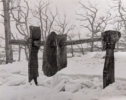 Black and white photo of a landscape in winter, the ground covered in snow. In the centre is an abstract sculpture, with thin sheets of what could be plaster or stone pierced by and hanging off a stout beam.