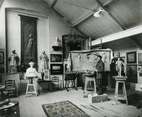Black and white photo of a young man in formal clothing, standing in a tidy art studio surrounded by figurative, relief and portrait sculpture.