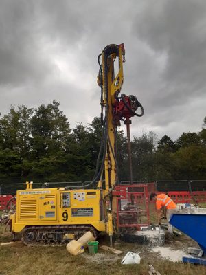 A yellow tracked vehicle with a drilling rig, with a man in an orange high-vis jacket working nearby.
