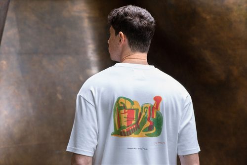 A white man with a freckled face and brown hair faces away from the camera. He is wearing a white t-shirt with an abstract design by Henry Moore of a reclining figure drawn in yellow, green and orange felt-tip pen. Behind the model is a bronze sculpture by Henry Moore.