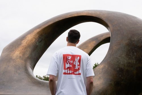 A white man with a freckled face and brown hair faces away from the camera. He is wearing a white t-shirt with an abstract design by Henry Moore of two white forms on a red background, drawn in felt-tip pen. Behind the model is the bronze sculpture 'Double Oval' by Henry Moore.