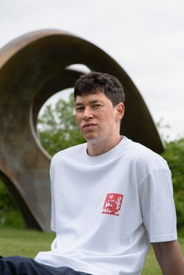 A white man with a freckled face and brown hair faces the camera. He is wearing a white t-shirt with a small abstract design by Henry Moore on the left breast, depicting two white forms on a red background, drawn in felt-tip pen. Behind the model is the bronze sculpture 'Double Oval' by Henry Moore.