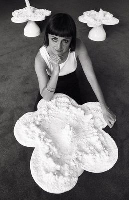 Black and white photo of a woman kneeling by a white sculpture of a 'flower' with five large petals. She is striking a pose with her hand on her chin. Two more similar sculptures are behind her.