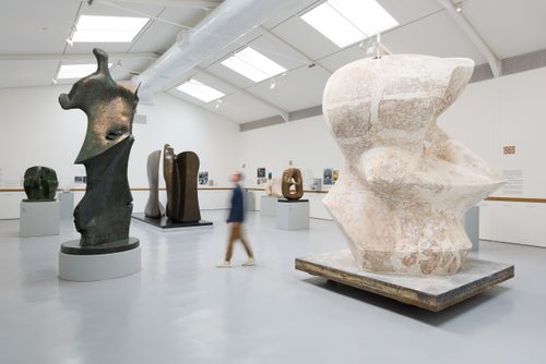A man walks between two sculptures by Henry Moore in a large gallery space: 'Large Spindle Piece' and 'Large Standing Figure: Knife Edge'.