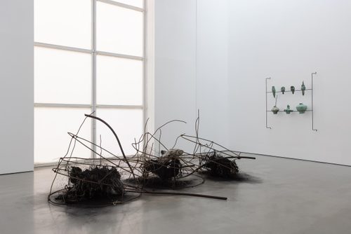 A gallery space with a sculpture made of burned material and rusted wire in the foreground, and a slender wire armature holding seven sculptures of roughly-made pots mounted on the wall behind.