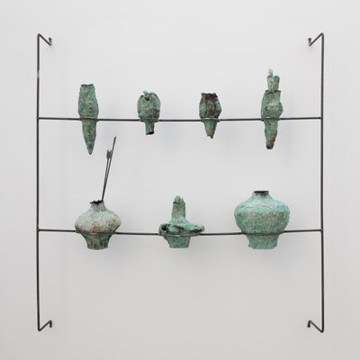 A slender wire armature mounted to a white wall, holding seven sculptures of roughly-made pots. They have a copper-green patina, and one has two arrows resting inside.