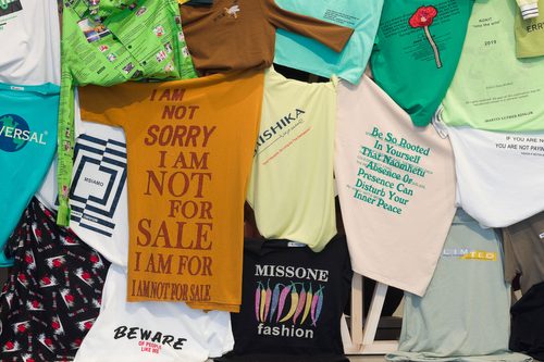 Close up photograph of several colourful t-shirts that make up Shanzhai Lyric's sculpture 'Incomplete Poem (hedge)'. The garments are part of a larger collection of bootleg slogan shirts, often featuring mistranslations. Some of the slogans on these t-shirts read: 'I AM NOT SORRY I AM NOT FOR SALE I AM FOR I AM NOT FOR SALE', 'BEWARE OF PEOPLE LIKE ME', 'Don't Muscle, As Long As The Demeanor', 'Be So Rooted In Yourself That Naomhetu Absence Or Presence Can Disturb Your Inner Peace' and 'MISSONE fashion'.