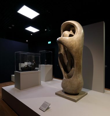 Gallery shot showing a large, upright stone sculpture by Henry Moore. The outside is smooth and roughly ovaloid, and the interior has been carved to make the outer shell appear like it is holding and protecting a smaller form within.