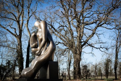 A tall, bronze sculpture depicting an abstracted mother and child is sited against a landscape of wintry trees with a bright blue sky behind