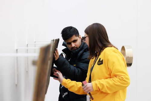 An exhibition guide in a bright yellow 'Beyond the Visual' top and a visitor are touching and listening to flat metal sculptures mounted to a gallery wall.