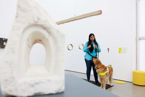 A visitor with a guide dog interacts with a sculpture, using a small hammer to tap a large ring suspended from a beam. In the foreground there is a stone sculpture with an opening carved in it in the shape of a window.