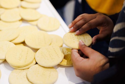 Close up of hands holding a plaster digestive biscuit with braille text and lettering reading 'comma'. Many more biscuits are on the table in front, featuring a variety of different words.