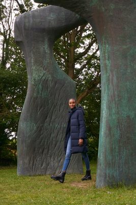 A model, wearing a long, navy puffer jacket, blue jeans and black boots, walks underneath Henry Moore's sculpture 'The Arch'.