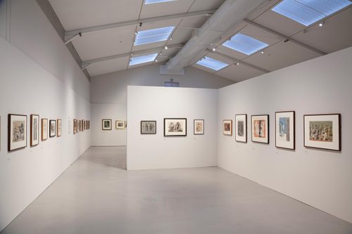 A high-ceilinged gallery space with a free-standing wall in the centre, on which several framed drawings are hung.