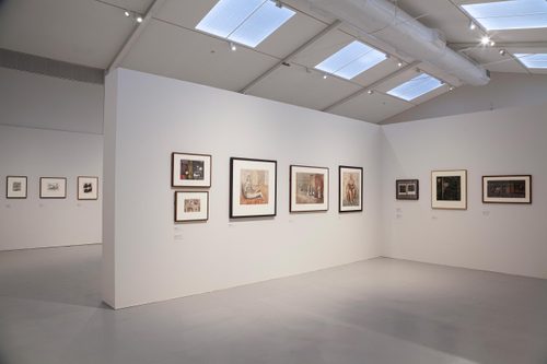 A high-ceilinged gallery space with a free-standing wall in the centre, on which several framed drawings are hung.