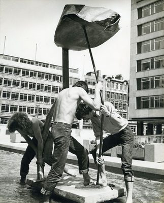 Black and white photo of three men, two of them shirtless, labouring to manoeuvre a large, umbrella-shaped metal sculpture into position.