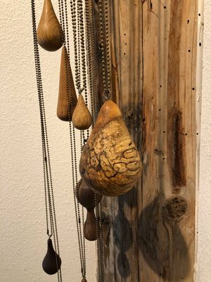 Wooden teardrop-shaped pendants hang from metal chains against a textured wooden post and white wall.