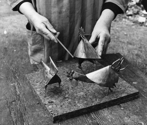 Close-in black and white photo of a pair of hands working on a sculpture of three small, stylised, metal sculptures of chicken-like birds.