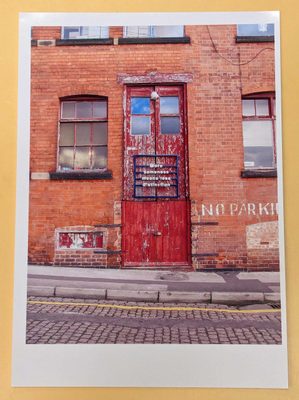 Printed photo of an industrial-looking red brick wall, with large, red wooden double doors, with the paint flaking off. A sign has been mounted to the door, which reads: 