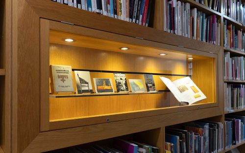 A display cabinet in the middle of a bookcase. It is displaying a series of five postcards by artist Joseph Beuys, and an open book.