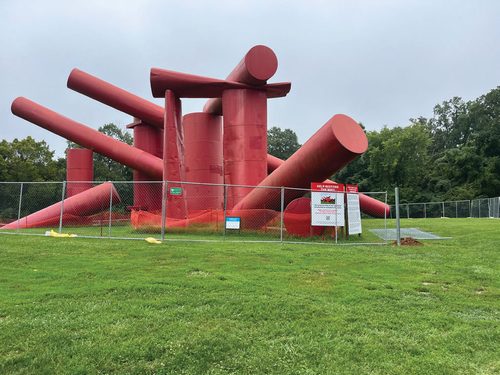 A large, badly-weathered piece of outdoor sculpture, made from metal cylinders painted red and piled together in a semi-haphazard way, behind a chain-link fence.