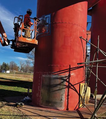 A man on a boom lift welding replacement sections of metal onto a large, red metal cylinder.