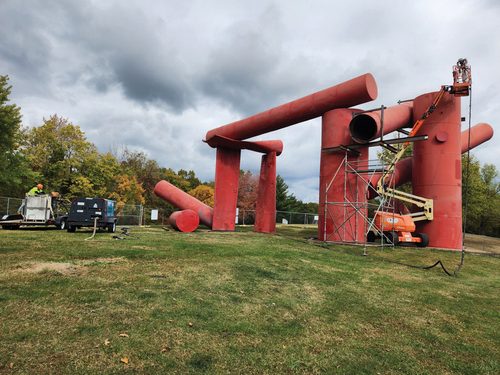 A boom lift and scaffolding enabling workmen to paint a large outdoor sculpture made up of several metal cylinders.