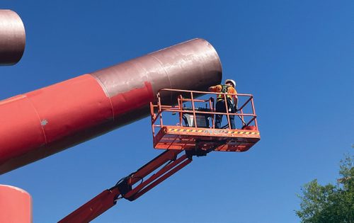 A man on a boom lift painting the end section of a large metal cylinder red, high in the sky.