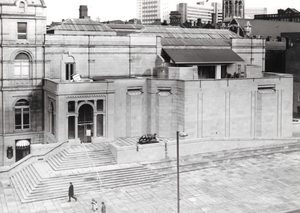 Black and white photo, taken from a high vantage point, of the front of Leeds Art gallery, a large stone building with steps and a ramp leading up an arched, glass-doored entrance.