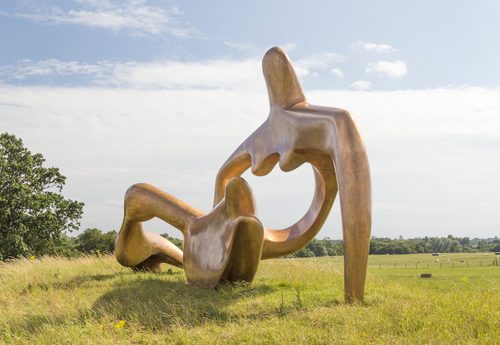 A huge abstract sculpture of a reclining figure, made in bronze. It has a large void in the centre of its torso. It is sited in a grassy field, with sheep in the distance behind.