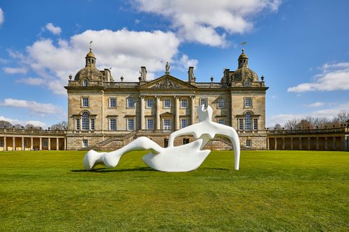 A large, white, fibreglass sculpture of a reclining figure in a green field in front of a large stone manor house.