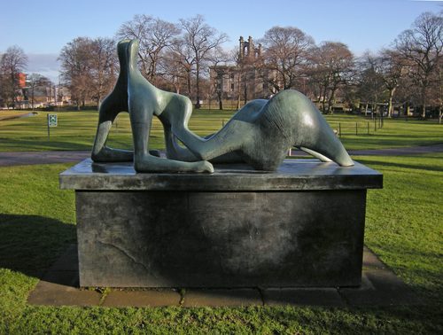 A colour photo showing a bronze reclining figure sculpture sited in a green park. In the background are stately stone buildings of the type you see in Scottish cities.