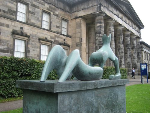 A colour photo showing a bronze abstract sculpture, of a reclining figure, sited outside an impressive Neo-classical building with columned entrance