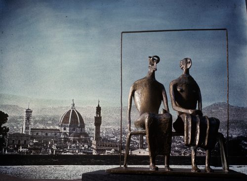 A small bronze model of 'King and Queen' by Henry Moore, photographed against an image of the Florence skyline, so that the model appears larger than it really is. The sculpture depicts two abstract figures seated on a bench, with a thin 'frame' around them.