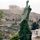 A tall bronze, abstract sculpture standing in the foreground with the famous Athens Acropolis visible on the hill behind.