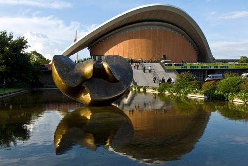 A colour photo of the dramatic Kongresshalle in Berlin, showing the curved roofline and water feature outside. Sited in the water is a huge bronze sculpture.