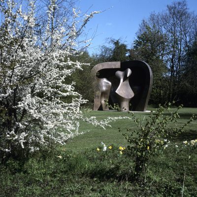 A hawthorn bush in bloom with daffodils scattered below. In the background is a huge bronze sculpture