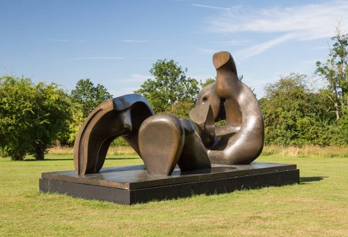 Photo of a large bronze sculpture. It is separated into three pieces, which together make an abstracted reclining figure. The sculpture is displayed on a plinth in a field of short grass, with trees and bushes behind.