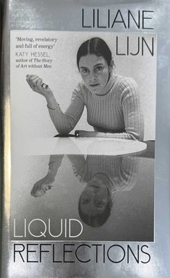 Cover of the book Liliane Lijn: Liquid Reflections, with a photo of the artist reflected in a table. Quote on the front of the book reads 'Moving, revelatory and full of energy' - Katy Hessel, author of The Story of Art without Men.